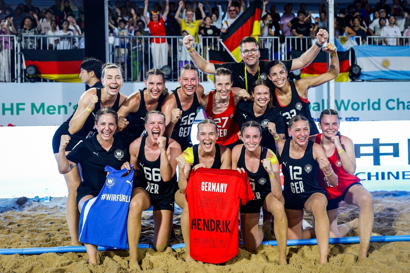 Das deutsche Beachhandball-Team jubelt gemeinsam nach dem Spiel und h&auml;lt Trikots mit den Aufschriften &bdquo;Germany&ldquo; und &bdquo;Hendrik&ldquo; in die Kamera.